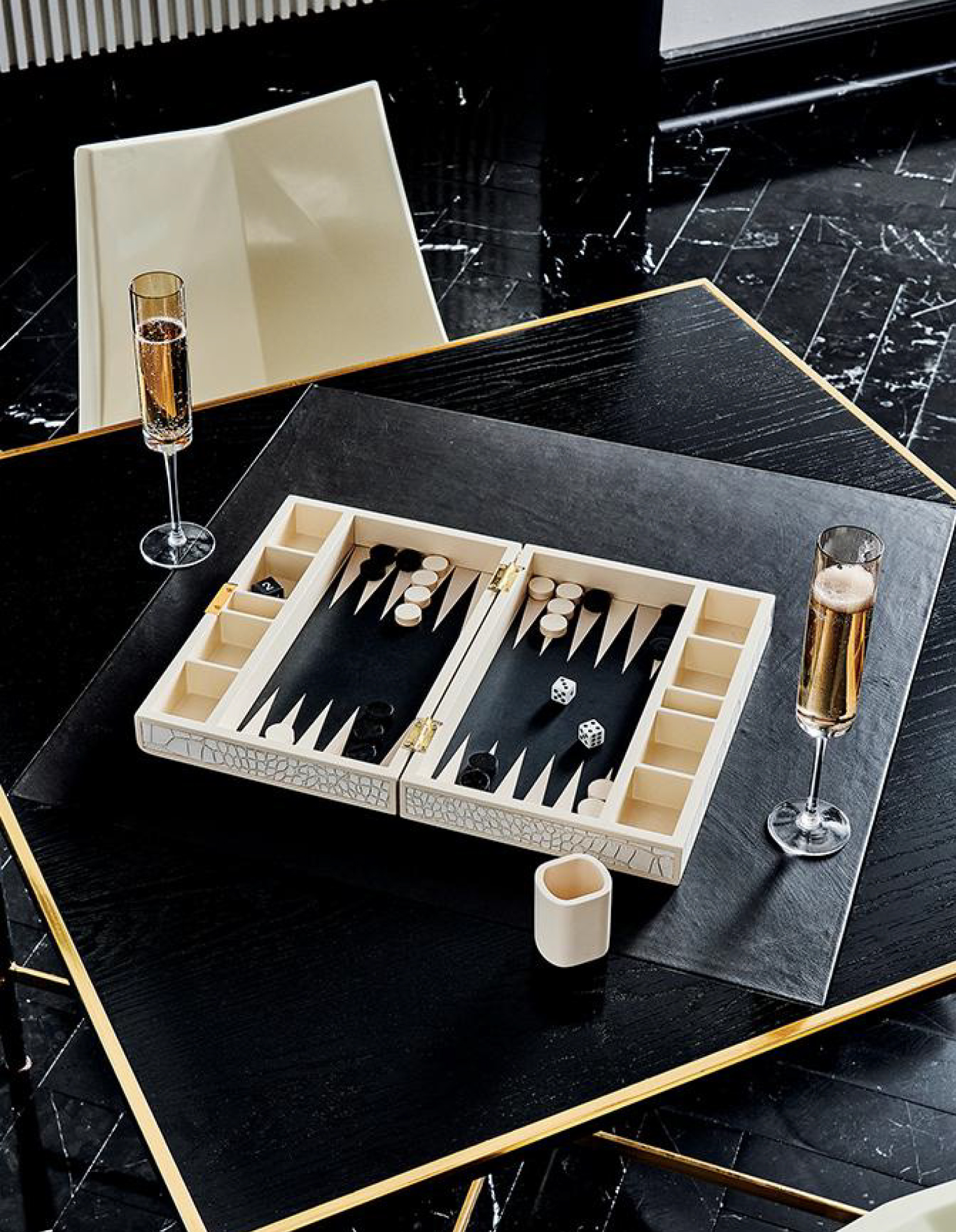 White ivory backgammon board with fake snakeskin cover beside two glasses of champaign on black wood table.